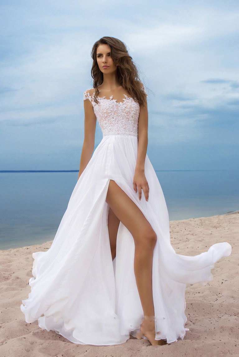 Woman in a white and pink wedding dress standing on a beach with blue sky and water in the background.